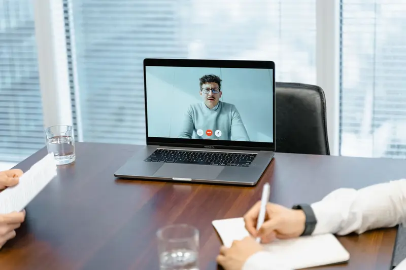 Videovergadering op een laptop aan een vergadertafel terwijl collega's notities nemen