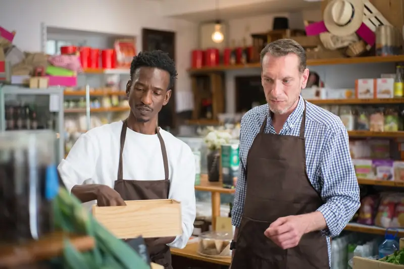 Twee medewerkers met schorten ordenen producten in de houten rekken van een buurtwinkel